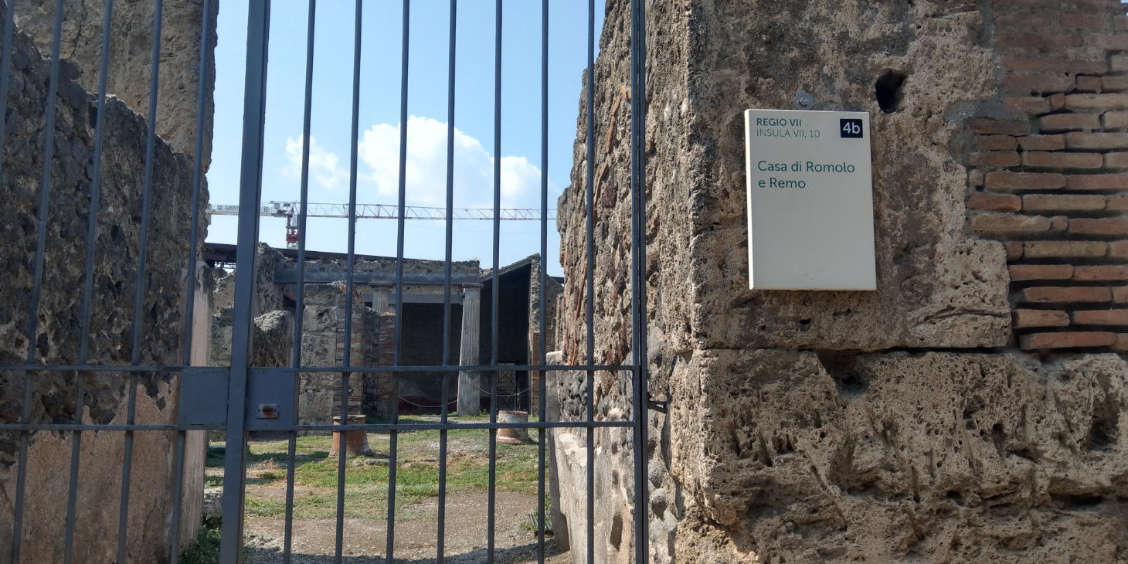 Vue rapprochée de l'entrée d'un site archéologique à Pompéi avec une grille en fer et une plaque pour la « Casa di Romolo e Remo ».