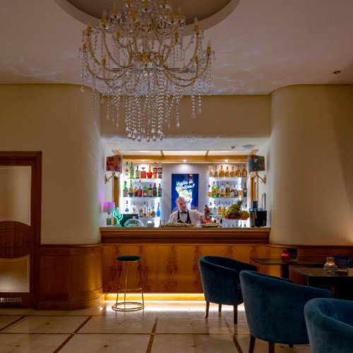Bar de l'hôtel avec lustre en cristal, fauteuils en velours bleu et comptoir en bois éclairé.