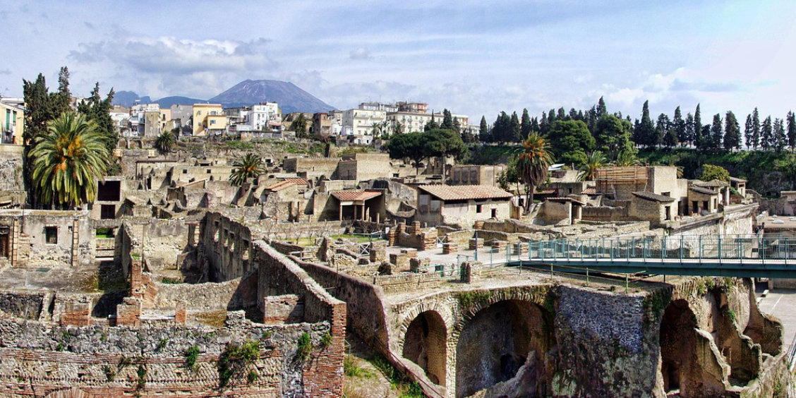 Vue d'ensemble des ruines antiques d'Herculanum avec des structures en pierre et brique, des arcades et le mont Vésuve se dressant à l'horizon derrière la ville moderne.