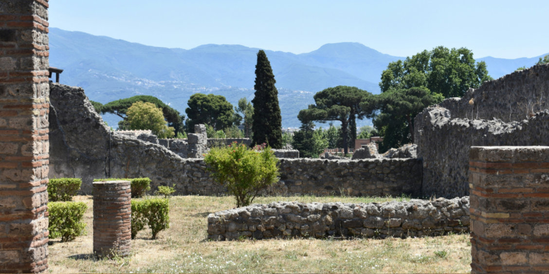 Ruines de la cité antique de Pompéi avec des murs en pierre, des arbustes et des arbres, devant un paysage de montagnes.