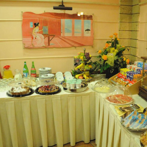 Buffet de petit-déjeuner garni de fruits frais, pâtisseries sous cloche, laitages et céréales, devant une peinture murale de style antique.