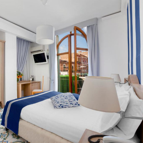 Chambre d'hôtel lumineuse avec un lit double aux touches bleues, un bureau en bois et une porte-fenêtre ouverte sur une terrasse.