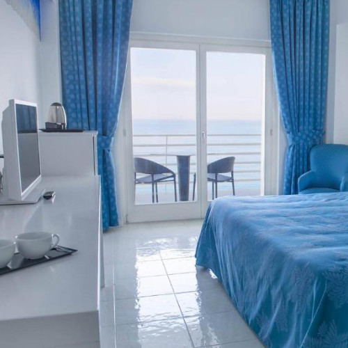 Intérieur d'une chambre d'hôtel dans les tons bleus et blancs avec une baie vitrée ouvrant sur un balcon face à l'horizon.