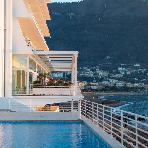 Vue de l'extérieur de l'hôtel moderne avec sa piscine en terrasse dominant la plage et les montagnes environnantes.