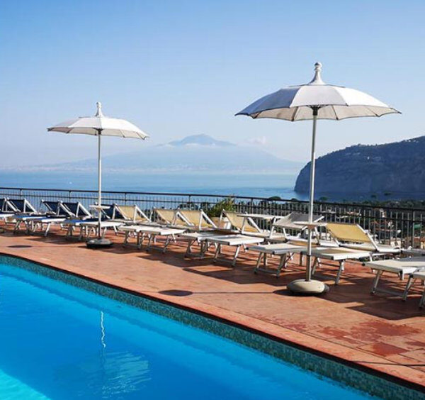 Piscine d’hôtel à Sorrente avec transats, parasols blancs et vue panoramique sur le golfe.