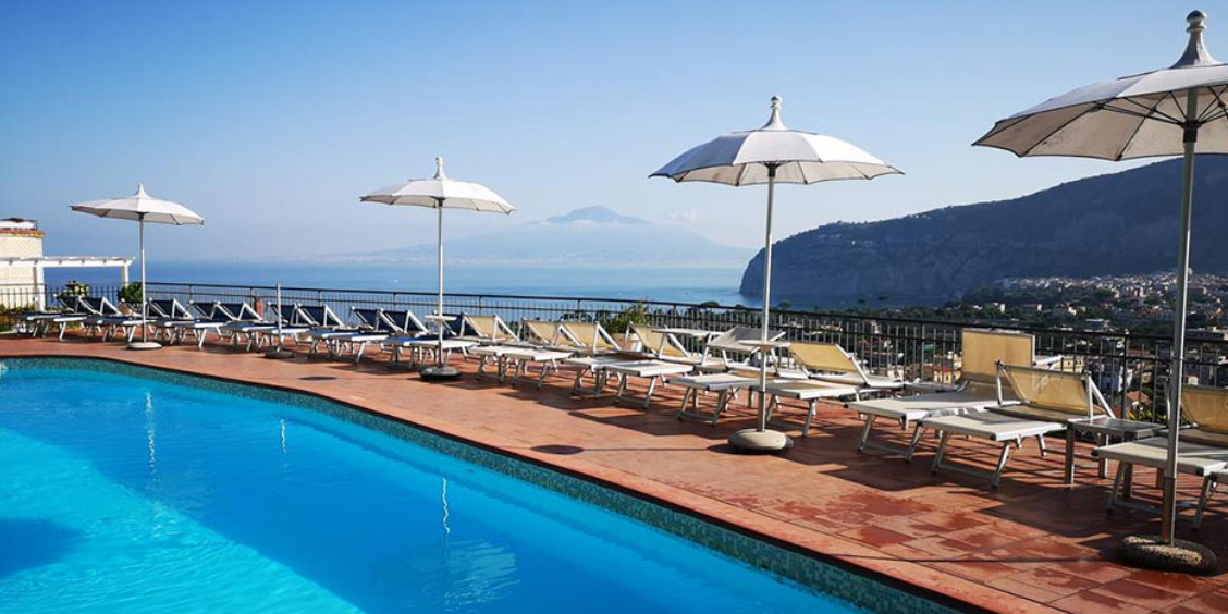 Piscine d’hôtel à Sorrente avec transats, parasols blancs et vue panoramique sur le golfe.