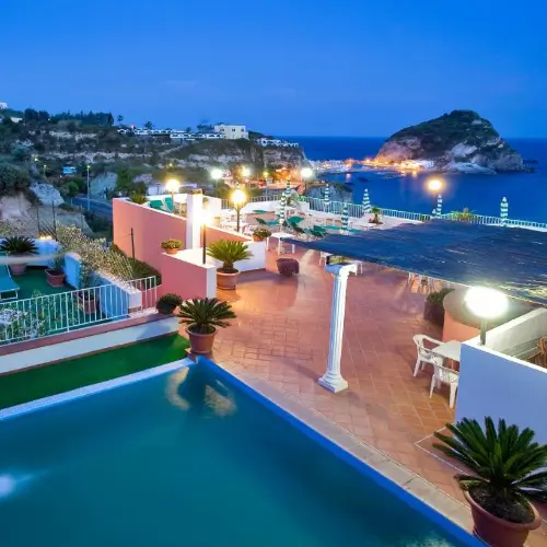 Piscine et terrasse de l'hôtel illuminées en soirée face à la baie de Sant'Angelo.