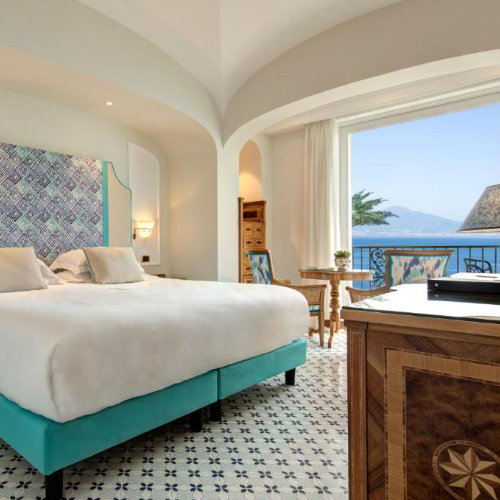 Chambre d'hôtel raffinée avec un lit double au sommier turquoise et un sol en mosaïque bleue, offrant une vue sur la mer et le Vésuve depuis le balcon.