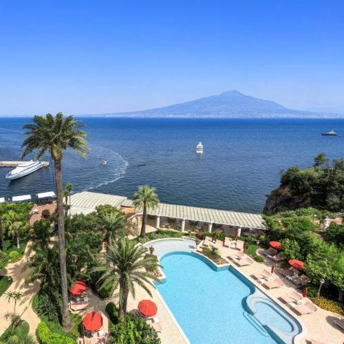 Vue aérienne du jardin luxuriant de l'hôtel avec sa piscine courbée et ses parasols rouges, face à l'immensité de la baie et à la silhouette du Vésuve.