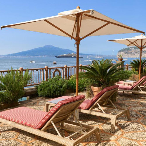 Terrasse ensoleillée équipée de transats rayés rouges et blancs sous de grands parasols, surplombant la mer parsemée de bateaux avec le Vésuve en arrière-plan.