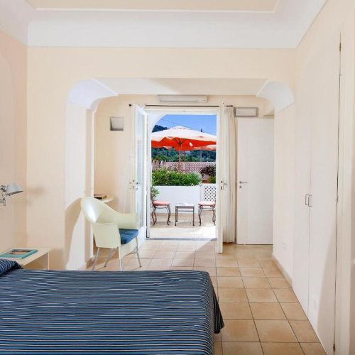 Chambre de l'Hôtel Senaria à Capri avec balcon ensoleillé, parasol orange et décoration épurée.