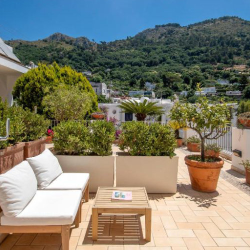 Terrasse de l'Hôtel Senaria à Capri avec canapés blancs, plantes en pot et vue sur la montagne verdoyante.