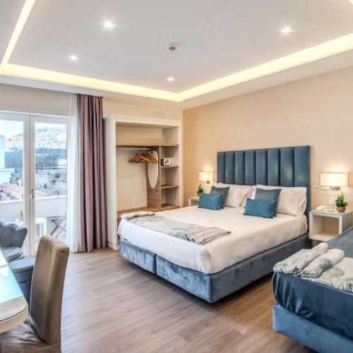 Chambre d'hôtel moderne et lumineuse avec un grand lit au sommier bleu, un placard ouvert en bois et une porte-fenêtre ouvrant sur un balcon.