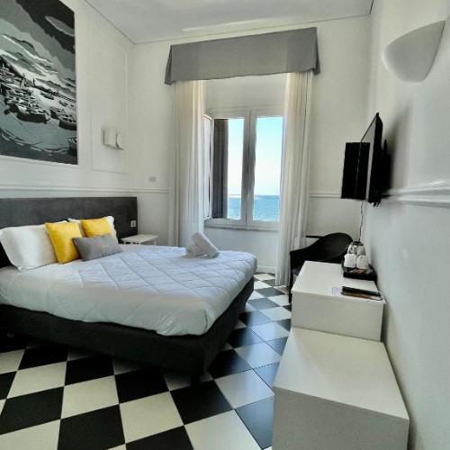Chambre d'hôtel lumineuse avec un sol en damier noir et blanc, un lit double orné de coussins jaunes et une fenêtre donnant sur la mer.