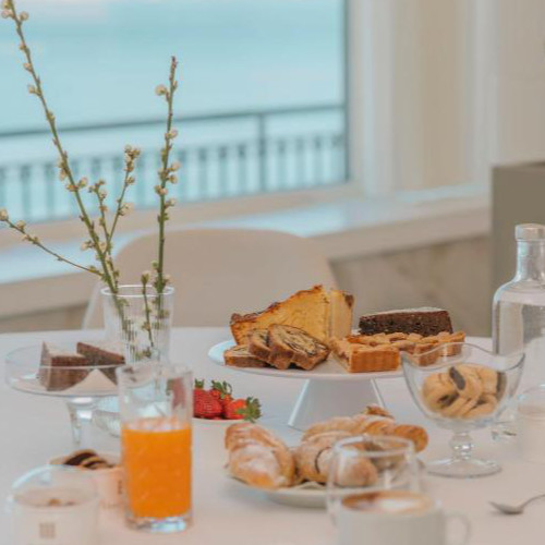 Petit-déjeuner copieux avec diverses pâtisseries, gâteaux et jus d'orange sur une table dressée face à une baie vitrée avec vue sur la mer.