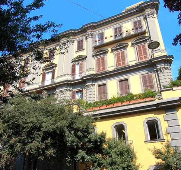 Façade d'un élégant palais napolitain aux tons jaunes et gris entouré de verdure.