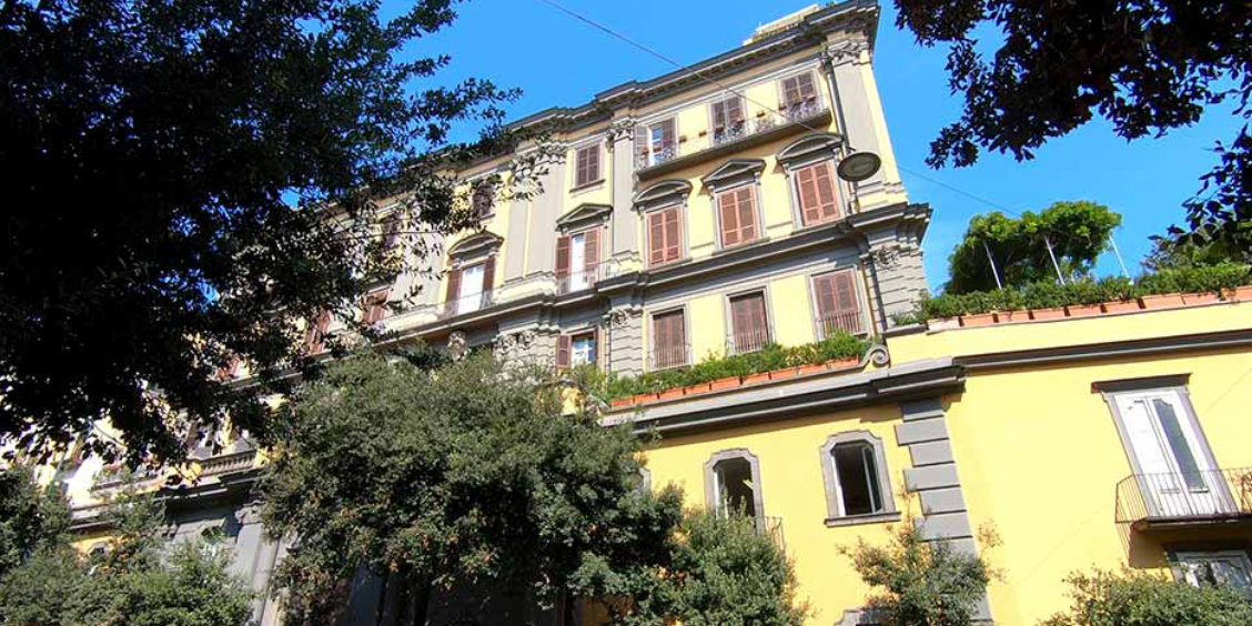 Façade d'un élégant palais napolitain aux tons jaunes et gris entouré de verdure.