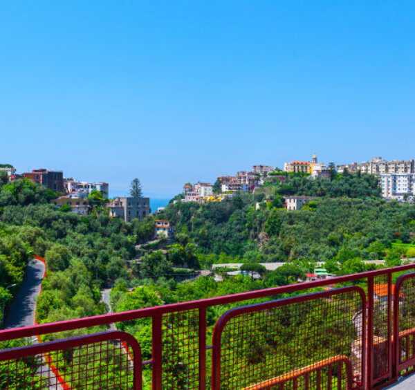 Gare de Sorrente avec horloge, passerelle rouge et vue sur les collines verdoyantes.