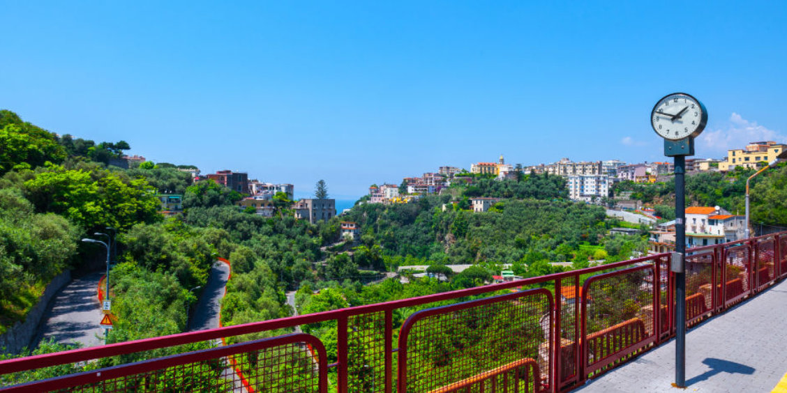 Gare de Sorrente avec horloge, passerelle rouge et vue sur les collines verdoyantes.
