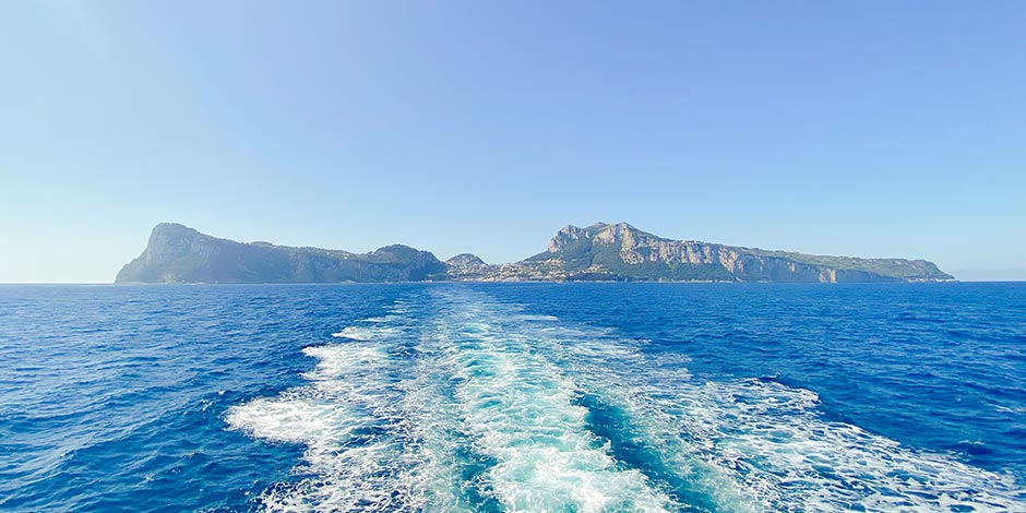 Île de Capri vue depuis la mer avec sillage de bateau sur l’eau bleue du golfe de Naples.