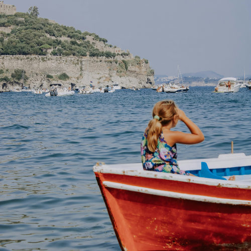 Une jeune fille sur un bateau rouge admirant la baie animée d'Ischia Porto.