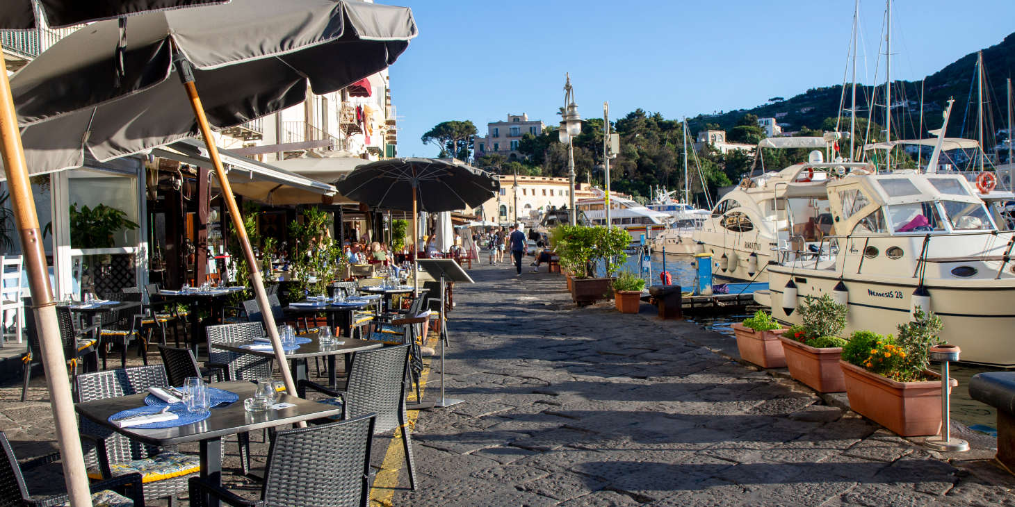 Terrasses de restaurants sur le front de mer d'Ischia Porto avec des bateaux amarrés.