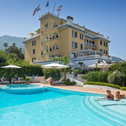 Façade d'un hôtel jaune élégant avec plusieurs étages et des drapeaux, surplombant une piscine animée avec des parasols blancs.