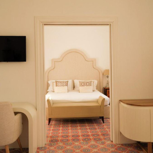 Chambre d'hôtel chic avec un lit double à tête de lit travaillée, vue à travers une porte sur une moquette aux motifs géométriques.