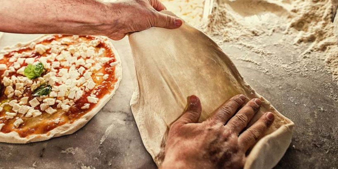 Gros plan sur les mains expertes d'un pizzaiolo étalant la pâte à pizza sur un comptoir en marbre, respectant le savoir-faire traditionnel reconnu.