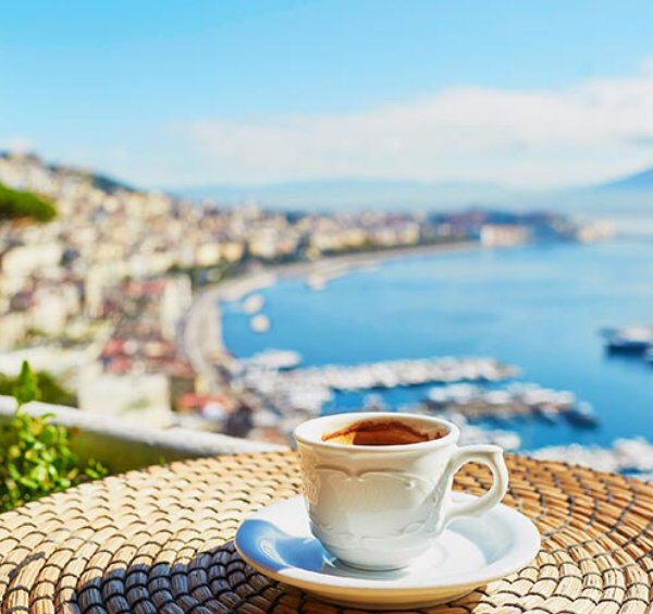 Tasse de café blanc sur une table tressée en terrasse, avec une vue panoramique sur la baie de Naples et le mont Vésuve à l'horizon.