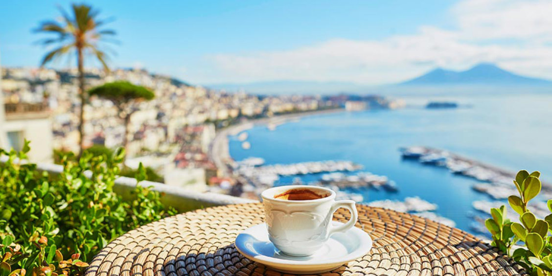 Tasse de café blanc sur une table tressée en terrasse, avec une vue panoramique sur la baie de Naples et le mont Vésuve à l'horizon.