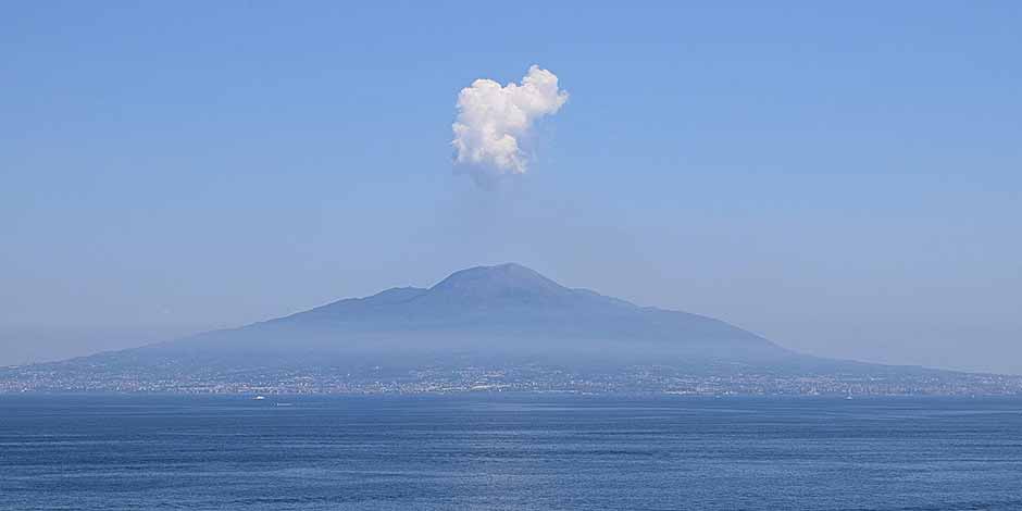 Le mont Vésuve se dressant au-delà de la mer sous un ciel bleu avec un unique nuage blanc au-dessus de son sommet.