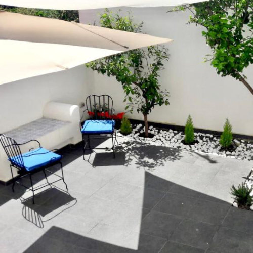 Terrasse moderne avec un sol en carrelage gris, des chaises en fer forgé aux coussins bleus et une zone de plantation avec des galets blancs.