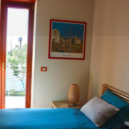 Chambre avec un lit aux draps bleus, une lampe de chevet tressée et un poster de Joan Miró au mur, à côté d'une porte-fenêtre.