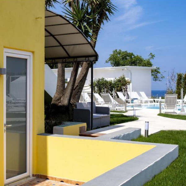 Façade jaune de l'hébergement avec une terrasse donnant sur l'espace piscine et la mer au loin.