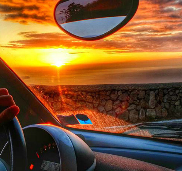 Vue depuis l'intérieur d'une voiture sur la Côte Amalfitaine avec coucher de soleil sur la mer, main sur le volant et muret en pierre.