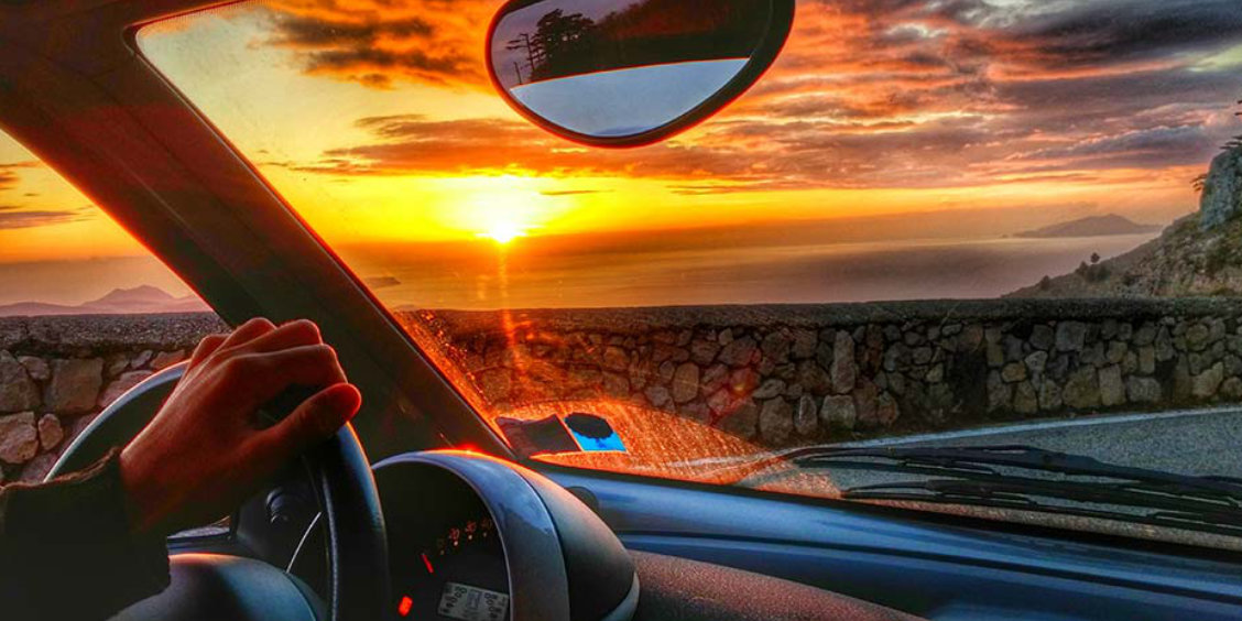 Vue depuis l'intérieur d'une voiture sur la Côte Amalfitaine avec coucher de soleil sur la mer, main sur le volant et muret en pierre.