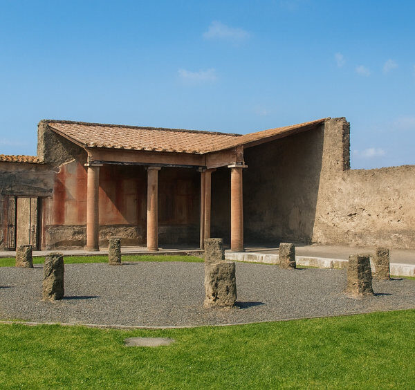 Ruines du Macellum à Pompéi montrant une structure circulaire entourée de pelouse verte et un bâtiment antique à colonnes sous un ciel bleu dégagé.