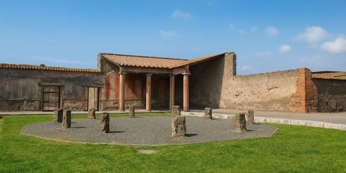 Ruines du Macellum à Pompéi montrant une structure circulaire entourée de pelouse verte et un bâtiment antique à colonnes sous un ciel bleu dégagé.