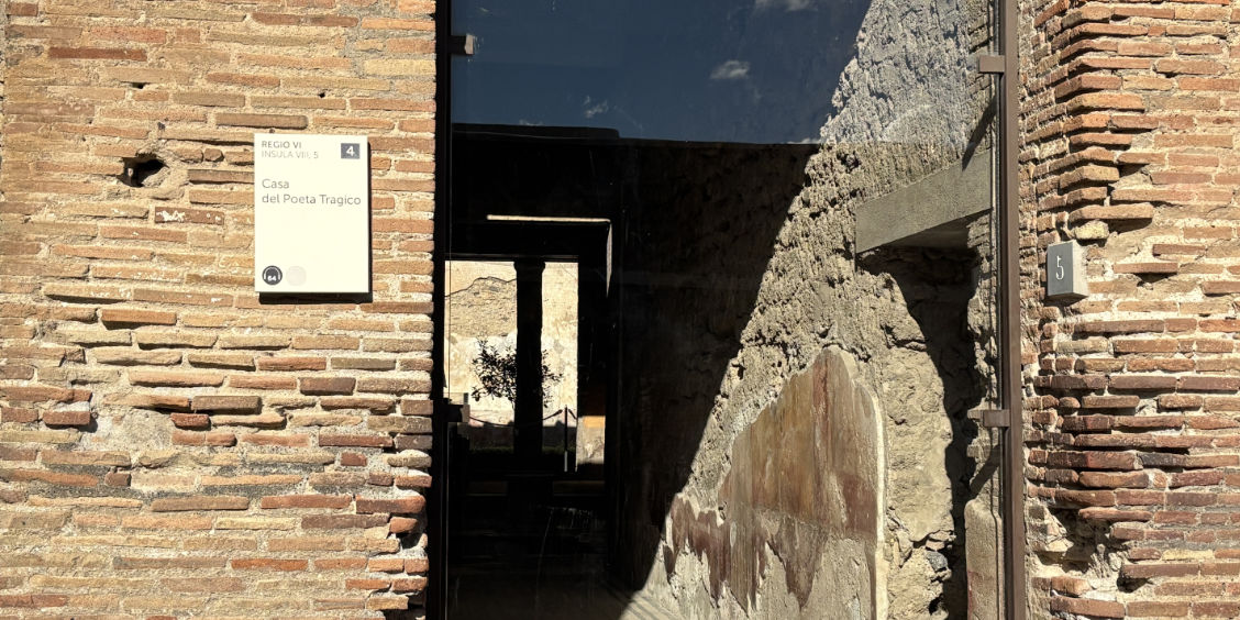 Entrée de la Maison du Poète tragique à Pompéi, montrant un mur de briques antiques et une plaque informative.