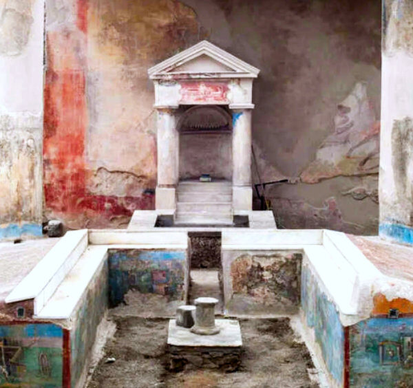 Triclinium d'été à la Maison de l'Éphèbe à Pompéi avec des banquettes ornées de fresques et un laraire en arrière-plan.