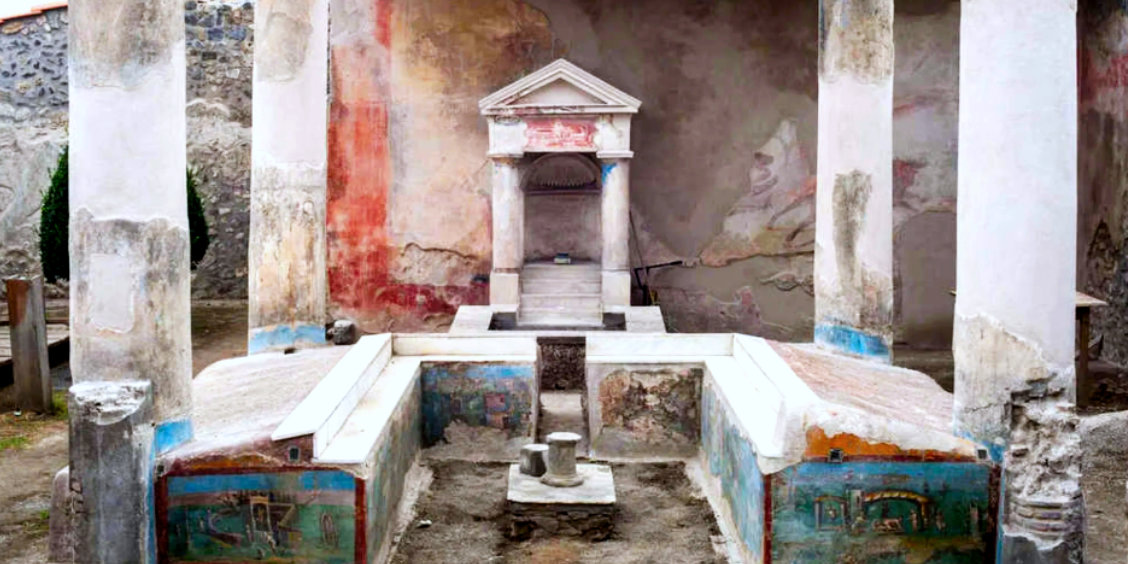 Triclinium d'été à la Maison de l'Éphèbe à Pompéi avec des banquettes ornées de fresques et un laraire en arrière-plan.