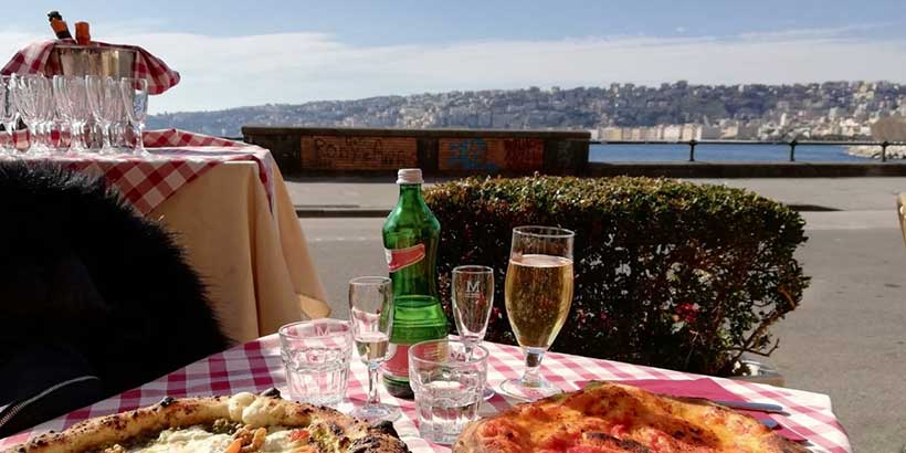 Table de restaurant à Naples avec pizzas et boissons face à la baie, vue panoramique sur la ville en arrière-plan.
