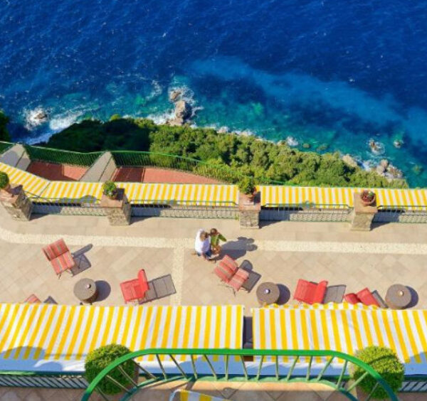 Terrasse d'hôtel à Anacapri avec auvents rayés jaunes, offrant une vue plongeante sur la mer et les falaises.