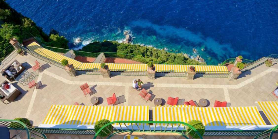 Terrasse d'hôtel à Anacapri avec auvents rayés jaunes, offrant une vue plongeante sur la mer et les falaises.