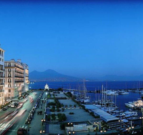 Vue du front de mer de Naples au coucher du soleil avec hôtels, port de plaisance et Vésuve en arrière-plan.