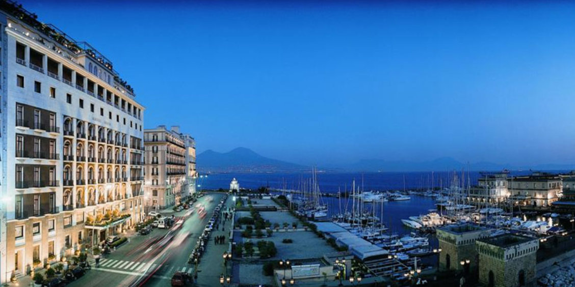 Vue du front de mer de Naples au coucher du soleil avec hôtels, port de plaisance et Vésuve en arrière-plan.
