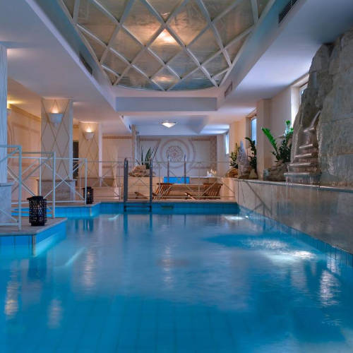 Piscine intérieure luxueuse avec cascade de pierre et plafond géométrique.
