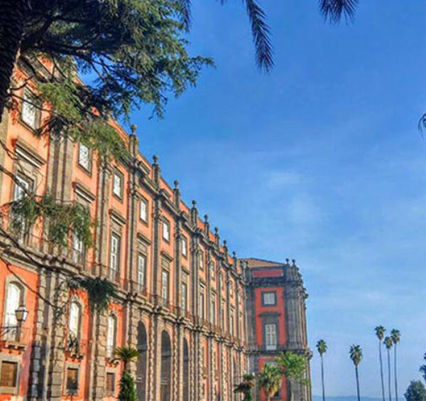Façade rouge néoclassique du musée national de Capodimonte à Naples, entourée de grands palmiers sous un ciel bleu.