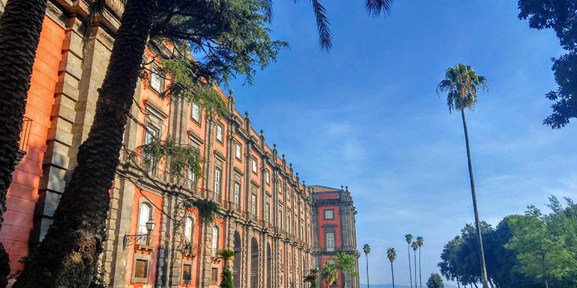 Façade rouge néoclassique du musée national de Capodimonte à Naples, entourée de grands palmiers sous un ciel bleu.